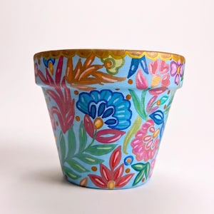 Könnte beinhalten: Ein hellblauer Terrakotta-Pflanztopf mit einem handgemalten Blumenmuster in Rosa, Rot, Dunkelblau und Grün. Der Rand des Topfes ist mit einer goldenen Bogenkante verziert, und die Außenseite zeigt verschiedene stilisierte Blumen und Blätter.