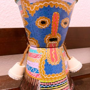 May include: Hand-painted terracotta flower pot with a whimsical design. The pot features a blue face with gold accents, pink nose, and black eyes. The lower section has colourful geometric patterns and small white bell-shaped accents. A unique decorative item.