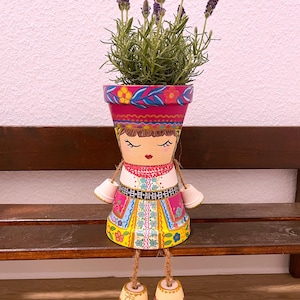 Puede incluir: Maceta de terracota pintada a mano con un diseño de arte popular. La maceta está decorada con una cara y un colorido estampado floral. Contiene una planta de lavanda con flores moradas. La maceta tiene piernas colgantes y está sobre un banco de madera.