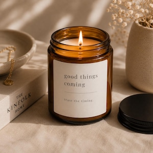 May include: A lit amber glass candle with a white label that reads "good things coming" and "trust the timing." The candle sits on a neutral surface next to a book and a small bowl with a gold necklace.