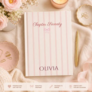 May include: A hardback journal with a white cover featuring vertical pink stripes. The text "Chapter Seventy" is printed in a script font above a small pink bow icon. The name "OLIVIA" is printed in a dark serif font near the bottom.
