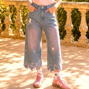 Op de afbeelding: Lichtblauwe wide-leg jeans van denim met een hoge taille, een kleine geborduurde kat bij de zak en een geschulpte zoom versierd met witte kattenomtrekken en sterrenmotieven, gecombineerd met een lichtroze T-shirt en roze sneakers.