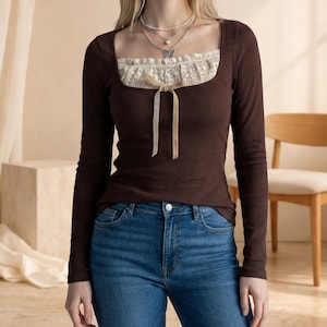 May include: A brown long-sleeve top with a square neckline and a cream-colored lace insert. The top features a ribbed texture and a beige bow detail at the neckline. The model is wearing blue jeans.