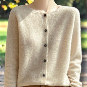 May include: A beige long-sleeved cardigan featuring a round neckline and a vertical row of five small, dark, circular buttons down the center front. The garment is crafted from a soft, textured knit fabric and is worn over brown trousers.