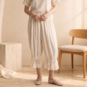 May include: A cream-coloured pajama set, featuring a short-sleeved top and cropped trousers with a drawstring waist and lace trim. The outfit is completed with beige flats. A wooden chair and cube are in the background. The outfit is made of a light fabric.
