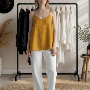 May include: A woman wearing a mustard yellow camisole top with thin straps and white pants. The top has a V-neck. A clothing rack with black and white shirts hangs in the background. A black hat and a small wooden stool are also visible.