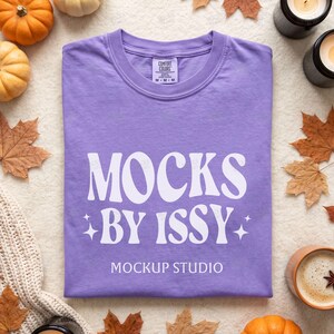 May include: A lavender short-sleeve t-shirt with a crew neckline. The front features white text that reads MOCKS BY ISSY with two four-pointed stars on either side, followed by the text MOCKUP STUDIO. The shirt has a Comfort Colors brand label.
