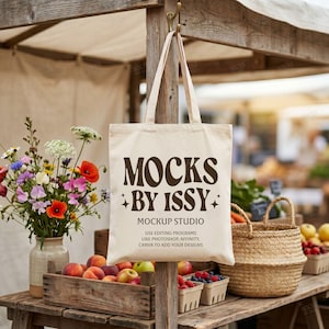 Peut inclure: Un sac fourre-tout en toile couleur crème suspendu à un poteau en bois. Le sac présente un texte marron foncé indiquant MOCKS BY ISSY MOCKUP STUDIO USE EDITING PROGRAMS LIKE PHOTOSHOP, AFFINITY, CANVA TO ADD YOUR DESIGNS avec des étoiles.