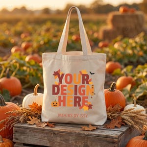 Könnte beinhalten: Eine beigefarbene Stofftasche mit dem Text "YOUR DESIGN HERE" in großen, herbstlich gefärbten Buchstaben, umgeben von Illustrationen aus Herbstblättern, einem kleinen Kürbisgesicht und einer Fledermaus, mit dem Aufdruck "MOCKS BY ISSY" darunter.
