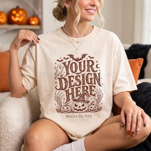 Könnte beinhalten: Ein cremefarbenes Kurzarm-T-Shirt mit einem braunen Grafikdruck mit dem Text "YOUR DESIGN HERE" und "Mocks by Issy". Das Design enthält Illustrationen eines Geistes, Fledermäusen, Skeletthänden und drei Kürbissen, die Hexenhüte tragen.