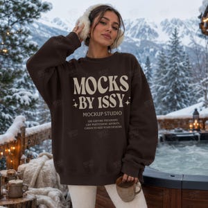 Könnte beinhalten: Ein dunkelbraunes Langarm-Sweatshirt mit cremefarbenem Text MOCKS BY ISSY MOCKUP STUDIO USE EDITING PROGRAMS LIKE PHOTOSHOP, AFFINITY, CANVA TO ADD YOUR DESIGNS und kleinen Sternsymbolen, getragen mit weißen Ohrenschützern und einer weißen Hose.