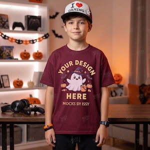 Può includere: Una t-shirt a maniche corte color bordeaux con la grafica di un fantasma bianco che indossa un cappello da strega viola, tiene in mano un secchiello a forma di zucca, circondato da zucche e pipistrelli, con il testo "YOUR DESIGN HERE" e "MOCKS BY ISSY" in giallo.