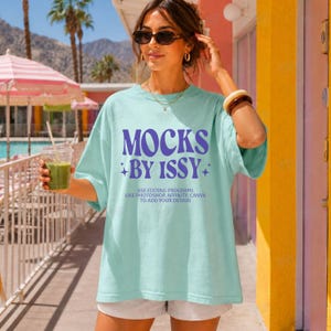 May include: A light blue t-shirt with the text "MOCKS BY ISSY" in purple. The shirt also includes the text "USE EDITING PROGRAMS LIKE PHOTOSHOP, AFFINITY, CANVA TO ADD YOUR DESIGN". The person is wearing sunglasses and holding a green drink.