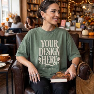 以下が含まれることがあります： セージグリーンの半袖Tシャツ。装飾的なセリフフォントでクリーム色の「YOUR DESIGN HERE」という文字がプリントされており、「HERE」の横には小さな葉のイラスト、その下には「Mocks by Issy」という小さな文字が配置されています。