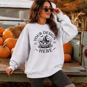 May include: A light gray crewneck sweatshirt featuring a black graphic print of a jack-o'-lantern wearing a witch hat, surrounded by bare trees and bats, with the text "YOUR DESIGN HERE" arched above and "EST. 2026" on the sides.