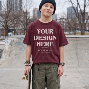 Può includere: Una t-shirt a maniche corte color bordeaux con il testo bianco YOUR DESIGN HERE, MOCKS BY ISSY e MOCKUP STUDIO, indossata con pantaloni cargo verde oliva, un berretto nero, un orologio da polso e uno skateboard in mano.