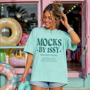 May include: A light blue t-shirt with the text "MOCKS BY ISSY" in dark teal. The shirt also has the text "MOCKUP STUDIO" and additional text below. The person is wearing denim shorts and has a bracelet on their wrist.