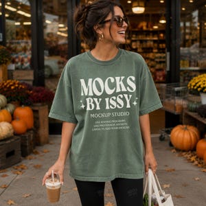 May include: A sage green short-sleeve t-shirt featuring the white text MOCKS BY ISSY MOCKUP STUDIO and instructions for use. The shirt has a crew neckline and a relaxed fit, displaying a vintage-style washed fabric texture on the front.