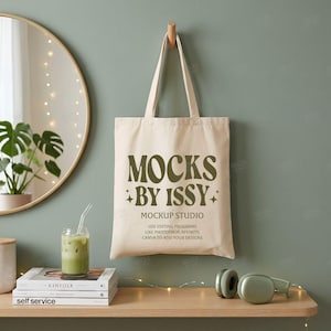 May include: A beige canvas tote bag hanging on a wall, featuring the dark green text MOCKS BY ISSY MOCKUP STUDIO USE EDITING PROGRAMS LIKE PHOTOSHOP, AFFINITY, CANVA TO ADD YOUR DESIGNS with small star icons on the front of the fabric bag.