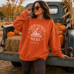 May include: An orange long-sleeve sweatshirt featuring a white graphic of a jack-o'-lantern wearing a witch hat, surrounded by tree branches. The text reads "YOUR DESIGN HERE", "EST. 2026", and "MOCKS BY ISSY" in a white, stylized font.