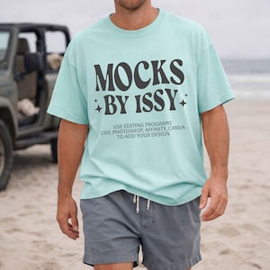 May include: A pale blue t-shirt with the words "MOCKS BY ISSY" in black, and the text "USE EDITING PROGRAMS LIKE PHOTOSHOP, AFFINITY, CANVA TO ADD YOUR DESIGN". The person is wearing grey shorts.