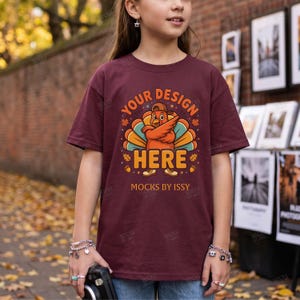 Può includere: Una t-shirt a maniche corte color bordeaux con la grafica di un tacchino dei cartoni animati che indossa un berretto da baseball, con il testo YOUR DESIGN HERE e MOCKS BY ISSY stampato sotto. Il tacchino arancione ha piume colorate.