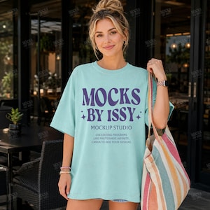 May include: A pale blue t-shirt with the text "MOCKS BY ISSY" in purple. The shirt also has the text "MOCKUP STUDIO" and additional text below. A striped tote bag is held in the right hand.