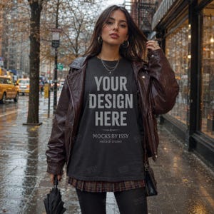 May include: A dark gray t-shirt featuring the white text YOUR DESIGN HERE and MOCKS BY ISSY MOCKUP STUDIO, worn under a brown leather jacket with a plaid skirt, black leggings, and a black shoulder bag, while holding a black umbrella.