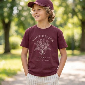Può includere: Una t-shirt a maniche corte color bordeaux con una grafica a linee bianche di un bouquet di fiori centrata sul petto. Il testo "YOUR DESIGN HERE" è stampato sopra i fiori e "MOCKS BY ISSY" è stampato sotto l'illustrazione floreale.
