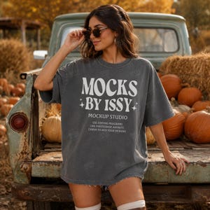 May include: A charcoal gray oversized t-shirt featuring white text that reads MOCKS BY ISSY MOCKUP STUDIO USE EDITING PROGRAMS LIKE PHOTOSHOP, AFFINITY, CANVA TO ADD YOUR DESIGNS with small star icons flanking the central text on the front.