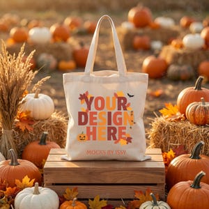 Peut inclure: Un sac fourre-tout en toile couleur crème portant le texte "YOUR DESIGN HERE" en grandes lettres colorées et grasses, entouré de motifs de feuilles d'automne, d'une petite citrouille et d'une chauve-souris. "MOCKS BY ISSY" est inscrit en dessous.