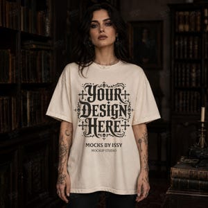 May include: Cream-colored t-shirt with the text "YOUR DESIGN HERE" in a gothic font. The shirt also features the text "MOCKS BY ISSY MOCKUP STUDIO". The t-shirt is a mockup, perfect for showcasing custom designs.
