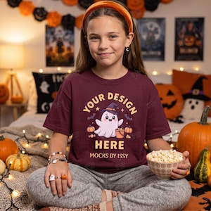 Può includere: Una t-shirt a maniche corte color bordeaux con la grafica di un fantasma bianco che indossa un cappello da strega viola, tiene in mano un secchiello a forma di zucca arancione, circondato da zucche, pipistrelli e caramelle, con il testo YOUR DESIGN HERE e MOCKS BY ISSY.