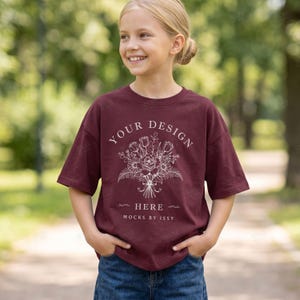 Può includere: Una t-shirt a maniche corte color bordeaux con una grafica a linee bianche raffigurante un bouquet di fiori al centro del petto. Il testo recita YOUR DESIGN HERE e MOCKS BY ISSY. La maglia ha un girocollo e una vestibilità ampia, abbinata a jeans blu.
