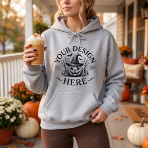 May include: A light gray hooded sweatshirt featuring a black graphic print of a jack-o'-lantern wearing a witch hat, surrounded by bare trees and bats, with the text "YOUR DESIGN HERE" and "EST. 2026" printed in a circular arrangement.