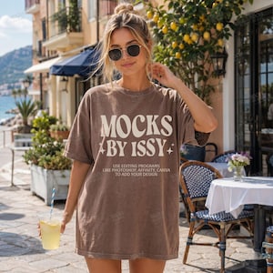 Op de afbeelding: Een bruin oversized t-shirt met de tekst "MOCKS BY ISSY" in het wit. Het shirt bevat ook de tekst "USE EDITING PROGRAMS LIKE PHOTOSHOP, AFFINITY, CANVA TO ADD YOUR DESIGN". Het shirt wordt gedragen door een persoon met een zonnebril.