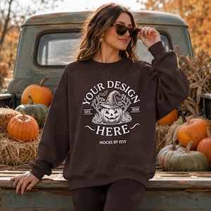 Peut inclure: Un sweat-shirt à manches longues marron foncé avec une impression graphique blanche représentant une citrouille d'Halloween portant un chapeau de sorcière, entourée d'arbres et de chauves-souris, avec le texte "YOUR DESIGN HERE", "EST. 2026" et "MOCKS BY ISSY".