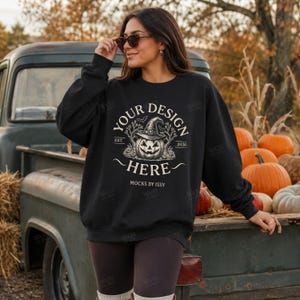 May include: A black long-sleeve sweatshirt featuring a white graphic print of a jack-o'-lantern wearing a witch hat, surrounded by trees and bats. The text reads "YOUR DESIGN HERE", "EST. 2026", and "MOCKS BY ISSY" in a circular layout.