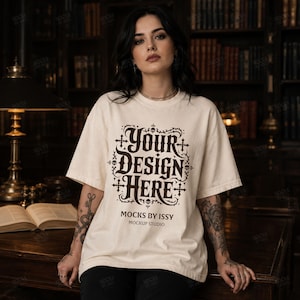 May include: Cream-colored t-shirt with the text "YOUR DESIGN HERE" in a dark brown, gothic-style font. The shirt also features the text "MOCKS BY ISSY" and "MOCKUP STUDIO" in a smaller font. The shirt is a mockup, perfect for showcasing designs.