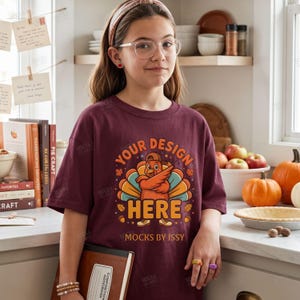 Può includere: Una t-shirt a maniche corte color bordeaux con la grafica di un tacchino che indossa un berretto da baseball ed esegue una posa dab. Il testo "YOUR DESIGN HERE" e "MOCKS BY ISSY" è in lettere arancioni e gialle, circondato da foglie autunnali e ghiande.