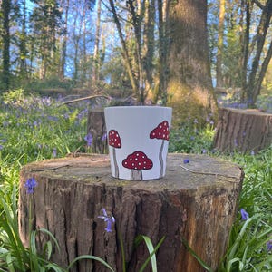May include: A white ceramic plant pot decorated with red mushrooms and white spots. The pot is cylindrical and stands on a tree stump in a natural outdoor setting, surrounded by trees and bluebells.