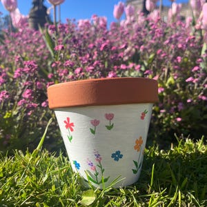 May include: A terracotta plant pot with a white painted exterior featuring small, hand-painted colourful flowers in shades of red, pink, orange, and blue with green stems and leaves. The pot has a wide, unpainted terracotta rim at the top.