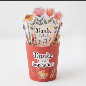 May include: A red cylindrical paper container featuring the text Danke FÜR DIE Inspiration, a paint palette, and a guitar. Inside are paper cutouts of flowers, two paintbrushes, a guitar, and a notepad with the text Danke FÜR DIE.