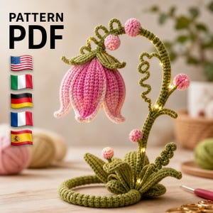 Könnte beinhalten: Eine gehäkelte Blumenlampe mit einem rosa und weißen Blütenkopf, einem grünen Stiel und rosa Beerenakzenten. Die Basis ist ein grüner Ring. Der Text "PATTERN PDF" befindet sich in der oberen linken Ecke. Die Lampe ist mit kleinen Lichtern beleuchtet.