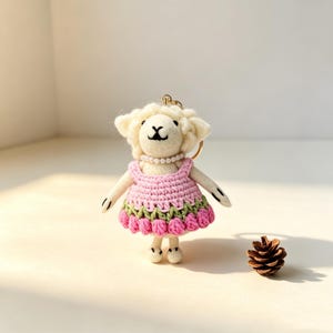 May include: A needle-felted sheep keychain featuring a cream-colored wool body, a pearl-bead necklace, and a crocheted dress with a pink bodice and a green and pink scalloped skirt. The sheep has black embroidered facial features and a gold ring.
