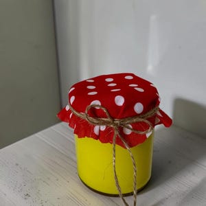 Puede incluir: Un tarro amarillo con una tapa de tela roja con lunares blancos atada con hilo de bramante. El tarro está lleno de una sustancia amarilla y se encuentra sobre una superficie de madera blanca. El tarro mide unos 10 cm de alto.