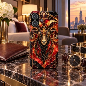 Puede incluir: Una funda para smartphone con una ilustración detallada de una cabeza de carnero con grandes cuernos curvados, en tonos rojo, negro y dorado. El diseño incluye un símbolo zodiacal de Aries en color oro en la parte superior y un patrón de vitral.