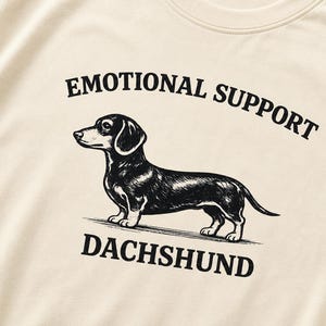 Può includere: Una t-shirt color crema con un'illustrazione a inchiostro nero di un bassotto in piedi di profilo. Sopra il cane, il testo recita EMOTIONAL SUPPORT, e sotto il cane, il testo recita DACHSHUND in un carattere nero, grassetto e senza grazie.