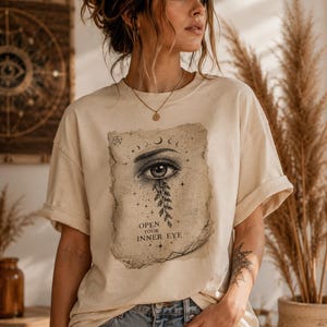 Könnte beinhalten: Cremefarbenes T-Shirt mit detaillierter Augengrafik und dem Text "OPEN YOUR INNER EYE". Das Design enthält himmlische Elemente und botanische Details. Das Shirt hat hochgekrempelte Ärmel und eine lockere Passform.