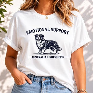 Può includere: Una t-shirt bianca a maniche corte con un'illustrazione grafica nera di un cane pastore australiano al centro. Sopra il cane, il testo recita EMOTIONAL SUPPORT, e sotto il cane, il testo recita AUSTRALIAN SHEPHERD.